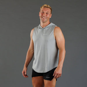 A smiling man wearing a light gray sleeveless hooded athletic shirt and black athletic shorts, standing against a solid dark gray studio background.