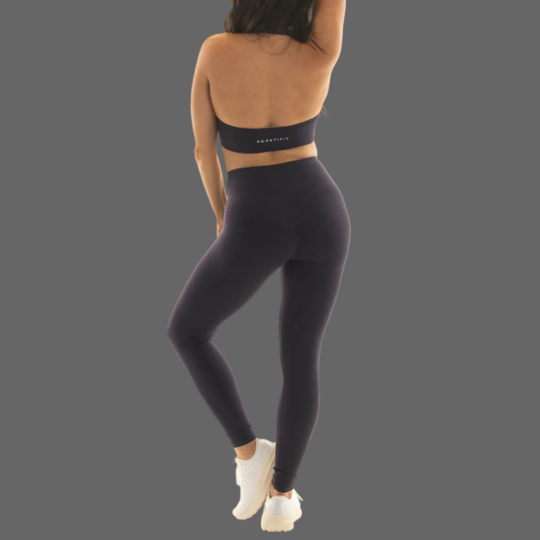 Rear view of a woman modeling dark charcoal gray high-waisted leggings and a matching sports bra against a solid gray background.