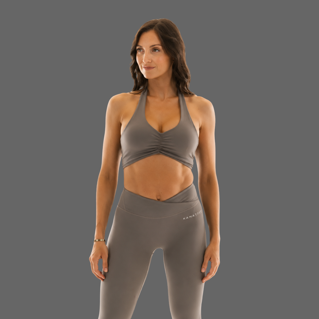A woman modeling a matching gray athletic set consisting of a ruched halter sports bra and high-waisted leggings, shown against a solid gray studio background.