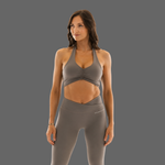 A woman modeling a matching gray athletic set consisting of a ruched halter sports bra and high-waisted leggings, shown against a solid gray studio background.