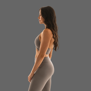 A woman in a side profile wearing a matching gray athletic set consisting of a sports bra and high-waisted leggings, set against a solid gray background.