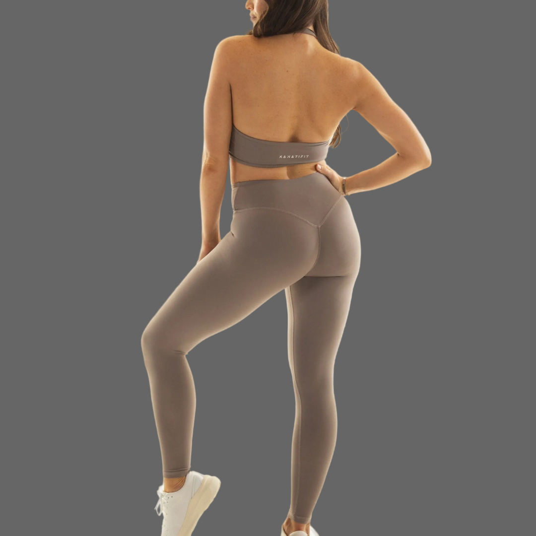 A woman shown from the back wearing a matching taupe-brown activewear set consisting of a sports bra and high-waisted leggings, paired with white sneakers, against a solid gray background.