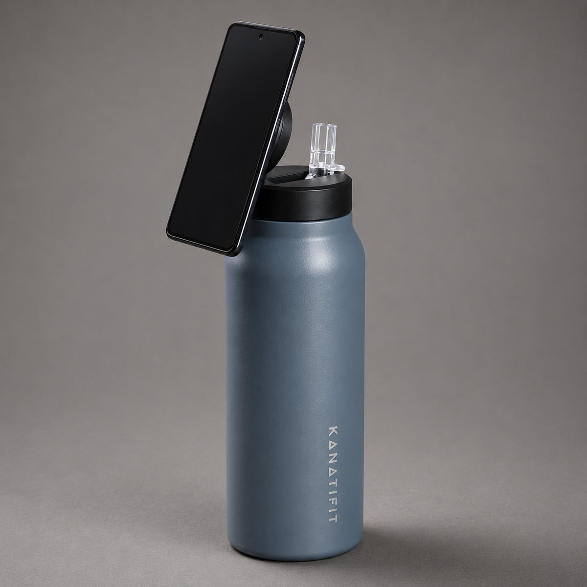 A matte slate insulated water bottle with a black lid and a black smartphone mounted on top, photographed on a smooth gray studio background. The brand name 'KANATIFIT' is printed vertically on the bottle.