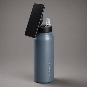 A matte slate insulated water bottle with a black lid and a black smartphone mounted on top, photographed on a smooth gray studio background. The brand name 'KANATIFIT' is printed vertically on the bottle.