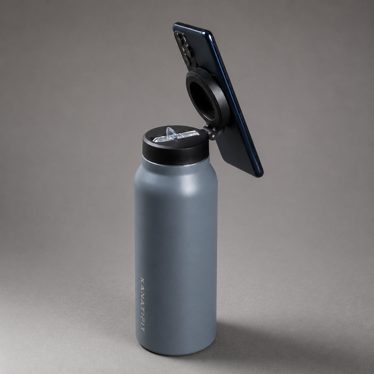 A matte gray insulated water bottle with a black flip-top lid, shown with a black smartphone mounted on the lid. The bottle has the brand name 'KANATIFIT' printed vertically near the base.