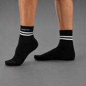 Black ankle socks with white double stripes and the brand name 'KANATIFIT' printed in white on the cuff, shown on a model's feet against a gray background.