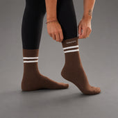A person wearing brown crew socks with two white horizontal stripes near the cuff, paired with black leggings, standing against a neutral gray studio background. The brand name 'KANATIFIT' is printed in white on the upper side of the right sock.