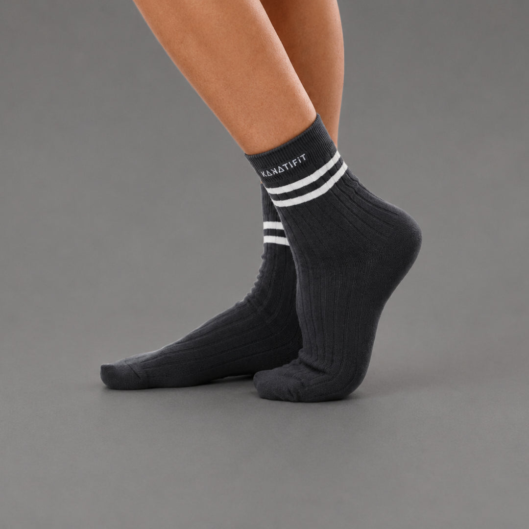 A pair of dark charcoal gray ankle socks with white stripes and the brand name 'KANATIFIT' printed in white on the cuff, shown on a model's feet against a solid gray background.