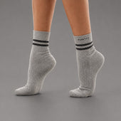 A pair of light gray heather ankle socks with two black stripes and the brand name 'KANATIFIT' printed on the cuff, shown on a model's feet against a solid gray background.