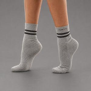 A pair of light gray heather ankle socks with two black stripes and the brand name 'KANATIFIT' printed on the cuff, shown on a model's feet against a solid gray background.