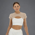 A woman modeling a beige short-sleeve crop top with a large chest cutout over a white sports bra, paired with white pants, against a solid gray studio background.