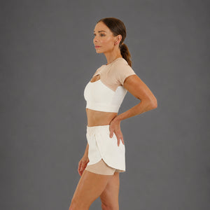 A woman modeling a matching beige and white activewear set, featuring a short-sleeve crop top with a chest cutout and high-waisted shorts, against a solid gray studio background.