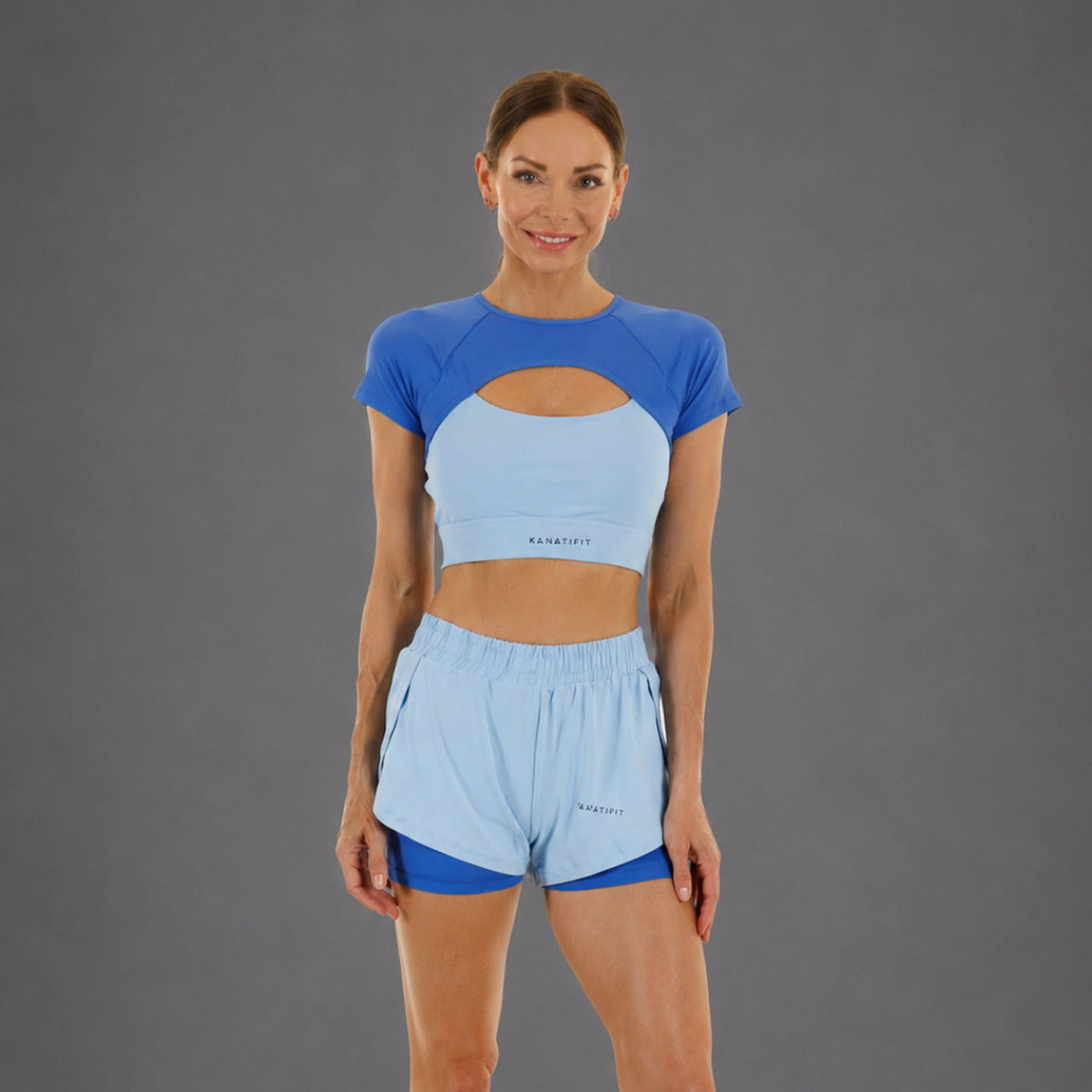 A woman modeling a matching athletic set consisting of a light blue and royal blue short-sleeve crop top and light blue running shorts, with the brand name 'KANATIFIT' visible on the front.