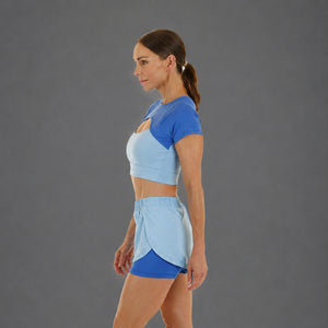 A woman in side profile wearing a two-tone athletic crop top and matching shorts in light blue and royal blue, photographed against a plain gray studio background.