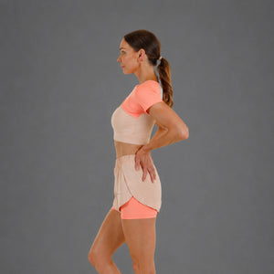 A woman in side profile wearing a peach and light beige athletic set, consisting of a short-sleeve crop top and matching wrap-style shorts, against a solid gray studio background.