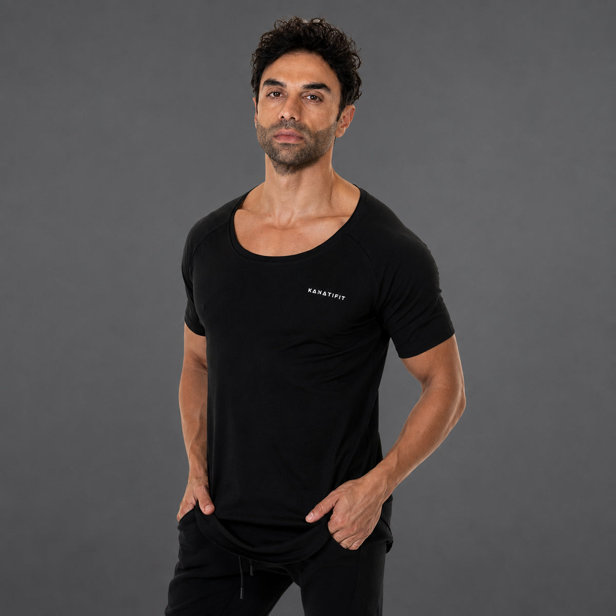 A man wearing a black scoop-neck athletic t-shirt with a small white 'KANATIFIT' logo on the left chest, posed against a solid dark gray studio background.