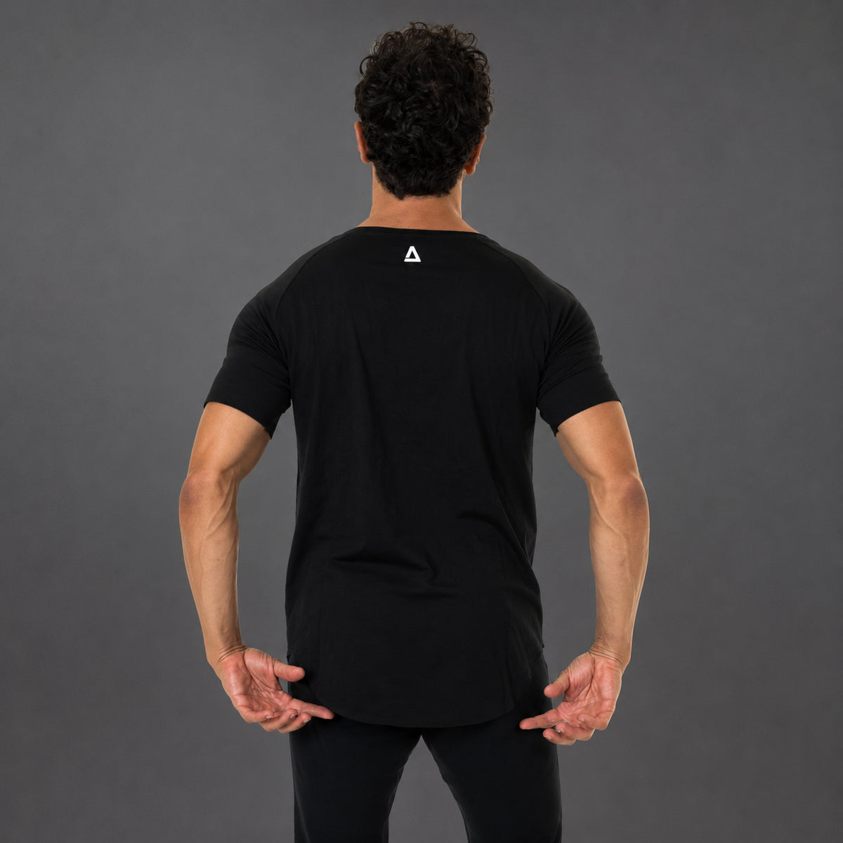 A man viewed from behind wearing a black short-sleeve athletic T-shirt with a small white geometric 'KANATIFIT' logo centered on the upper back, paired with black pants, against a dark gray studio background.