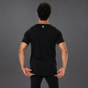 A man viewed from behind wearing a black short-sleeve athletic T-shirt with a small white geometric 'KANATIFIT' logo centered on the upper back, paired with black pants, against a dark gray studio background.