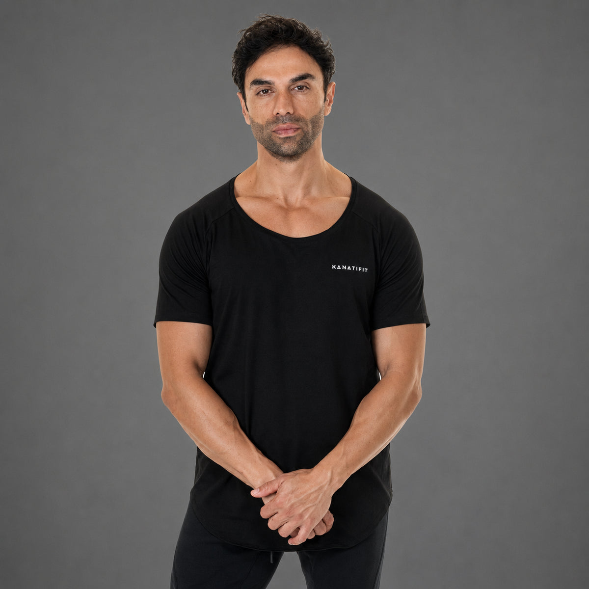 A man wearing a black short-sleeve athletic t-shirt with a wide scoop neckline and a small white 'KANATIFIT' logo on the left chest, standing against a solid gray studio background.