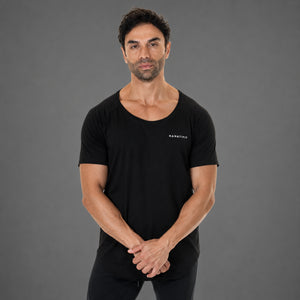 A man wearing a black short-sleeve athletic t-shirt with a wide scoop neckline and a small white 'KANATIFIT' logo on the left chest, standing against a solid gray studio background.