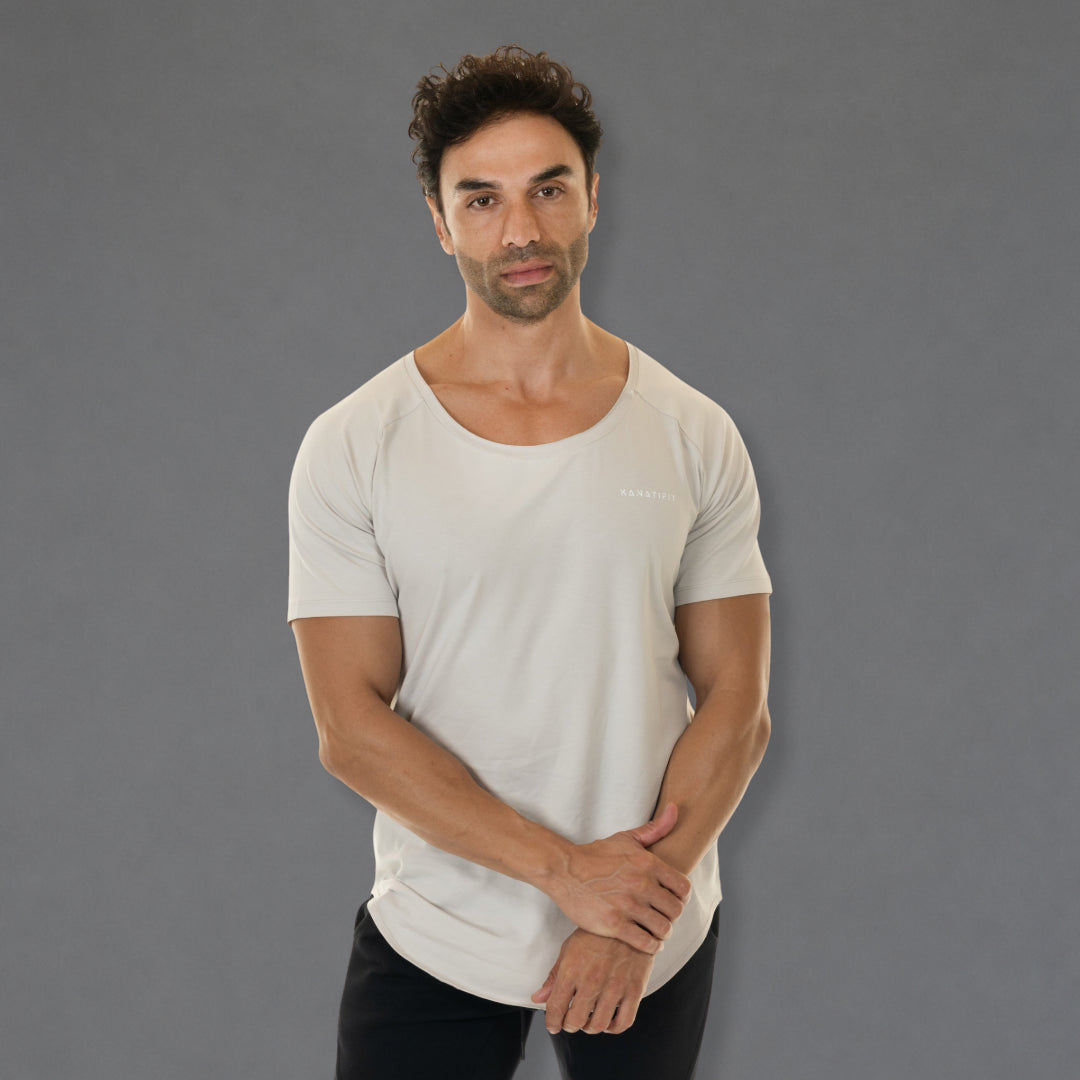 A man wearing a light beige scoop-neck athletic t-shirt with a small white logo on the left chest, paired with black pants, standing against a solid gray studio background.