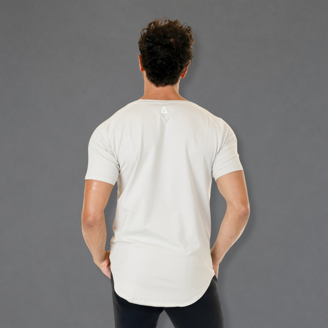 A man viewed from behind wearing a white short-sleeve athletic t-shirt with a curved hem and a small geometric logo on the upper back, paired with black pants, against a solid gray background.