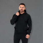 A man wearing a black pullover hoodie and matching black sweatpants, standing against a solid gray studio background.