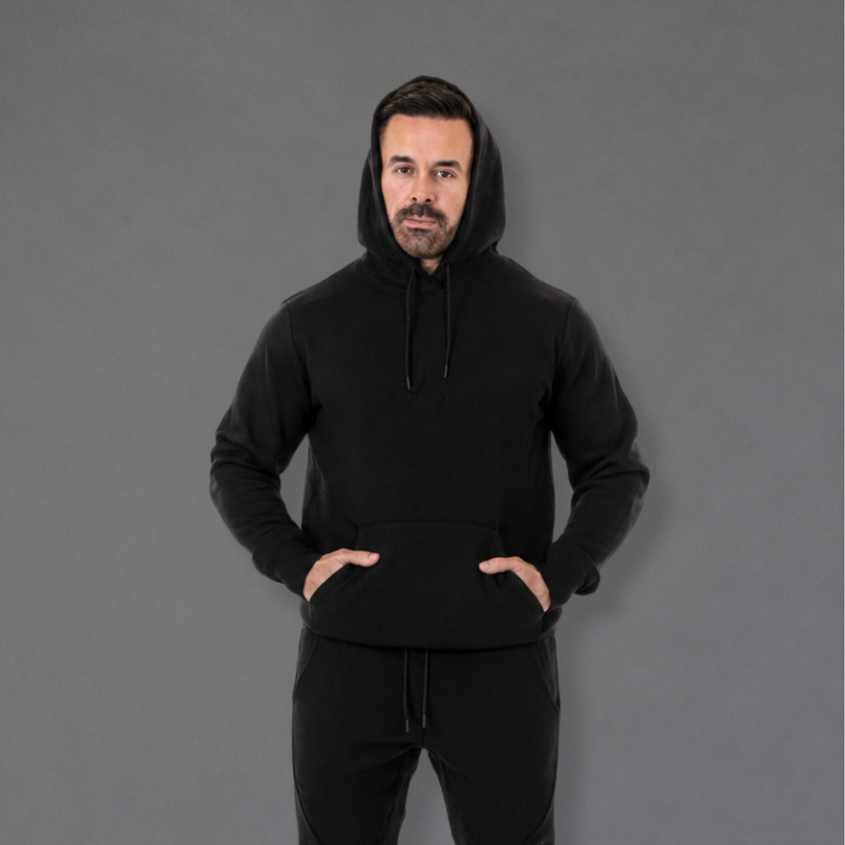 A man wearing a black pullover hoodie with a drawstring hood and front kangaroo pocket, paired with black sweatpants, standing against a solid gray studio background.