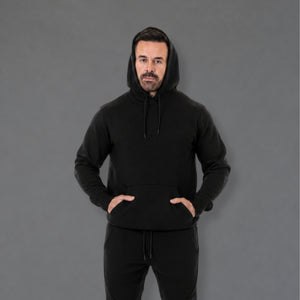 A man wearing a black pullover hoodie with a drawstring hood and front kangaroo pocket, paired with black sweatpants, standing against a solid gray studio background.