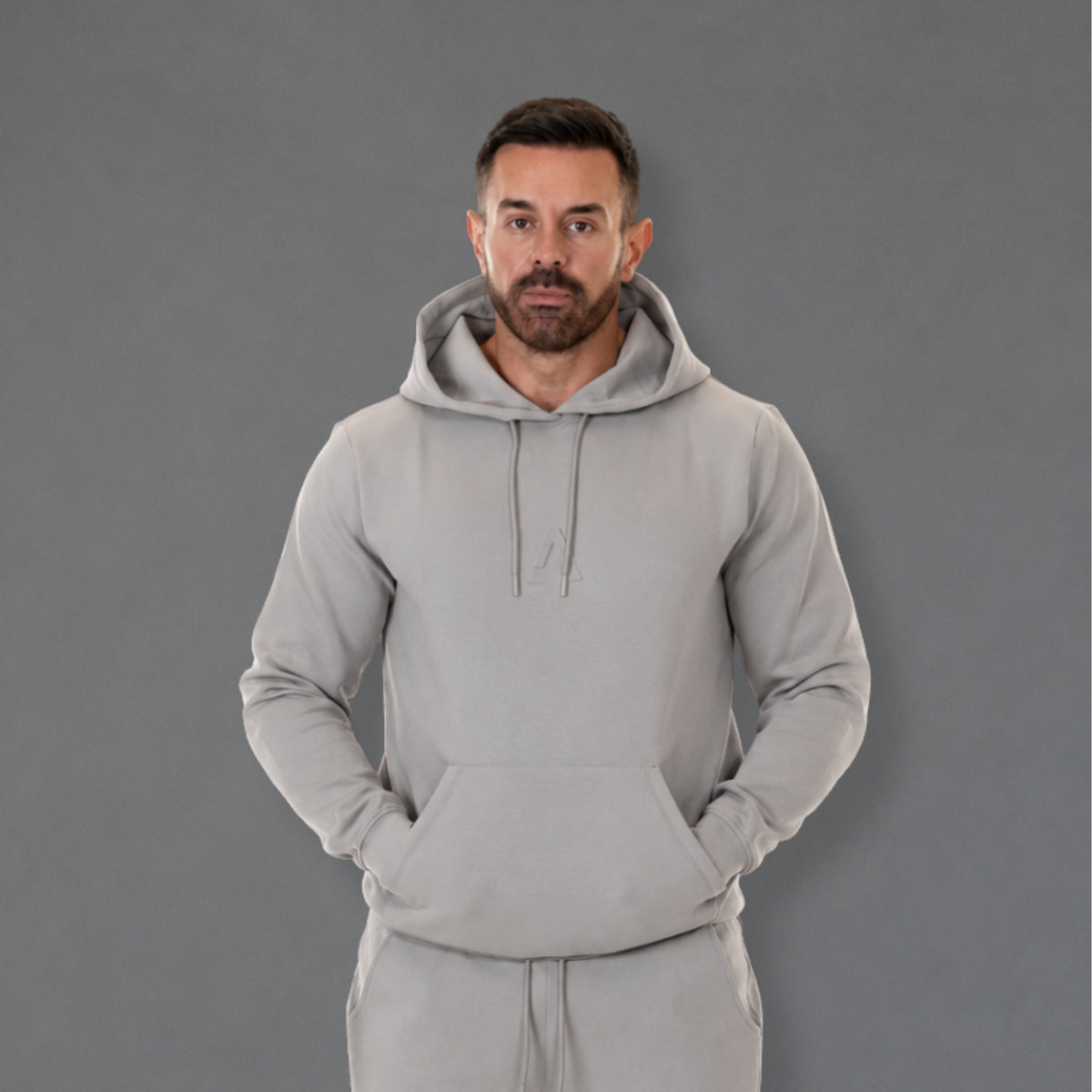A man wearing a light gray pullover hoodie and matching sweatpants, standing against a solid gray studio background.