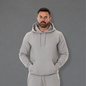 A man wearing a light gray pullover hoodie and matching sweatpants, standing against a solid gray studio background.