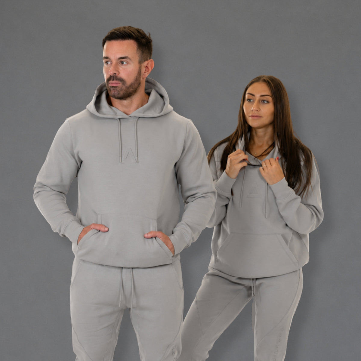 A man and a woman modeling matching light gray hoodies and sweatpants sets against a solid gray studio background.