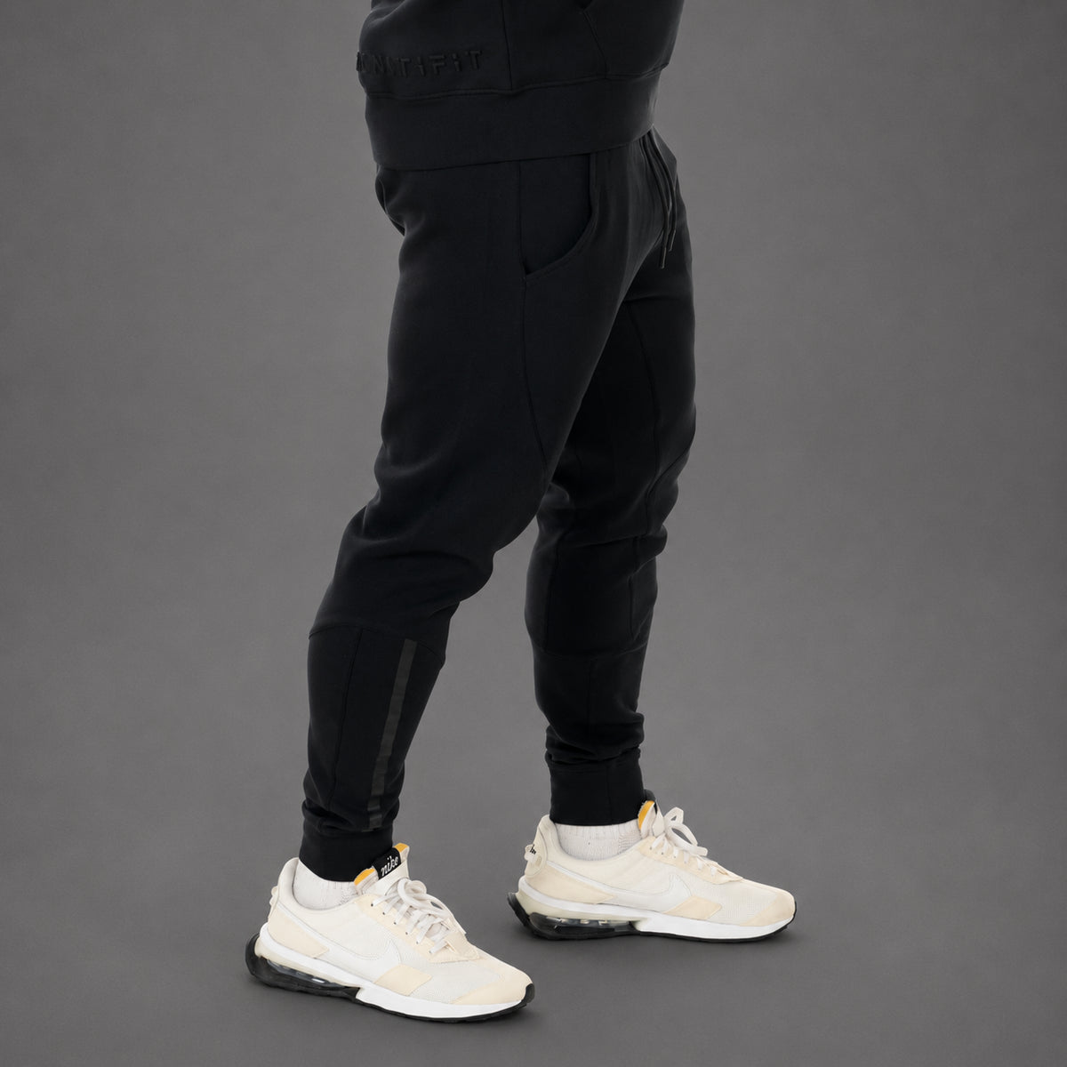 A person wearing black jogger-style pants with side compression panels and white sneakers, shown from the waist down against a dark gray studio background.