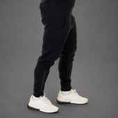 A person wearing black jogger-style pants with side compression panels and white sneakers, shown from the waist down against a dark gray studio background.
