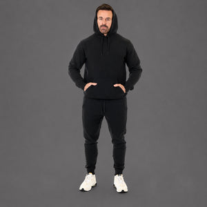 A man wearing a black pullover hoodie and matching black jogger pants, standing against a solid dark gray studio background. He is wearing white sneakers.