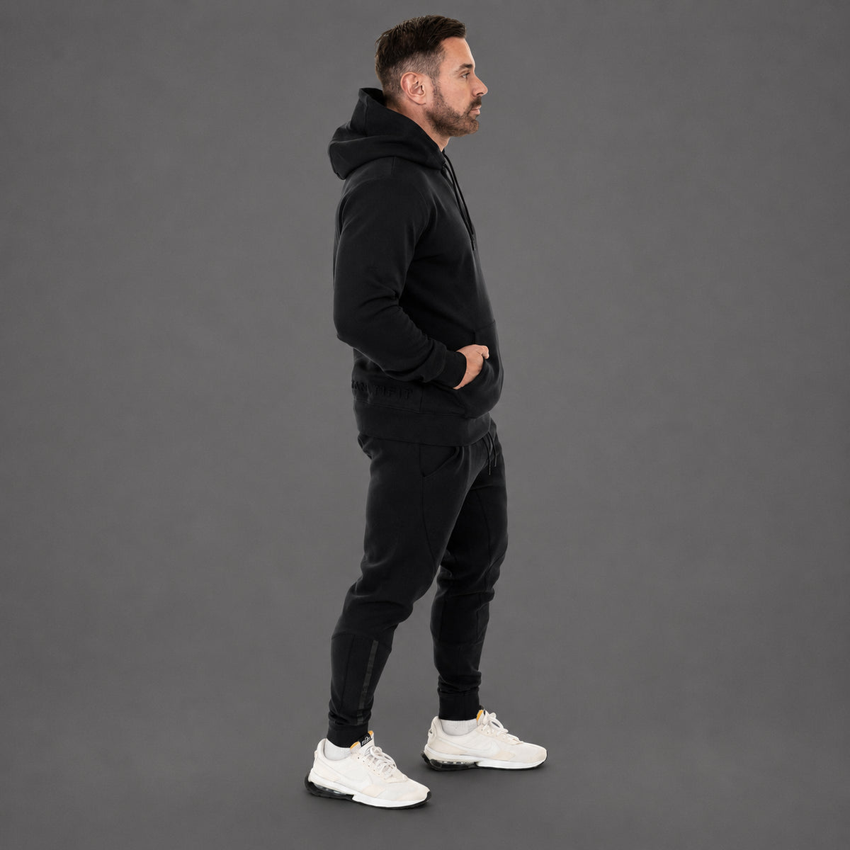 A man in a black hooded sweatshirt and black jogger pants stands in profile against a solid gray studio background, wearing white sneakers.