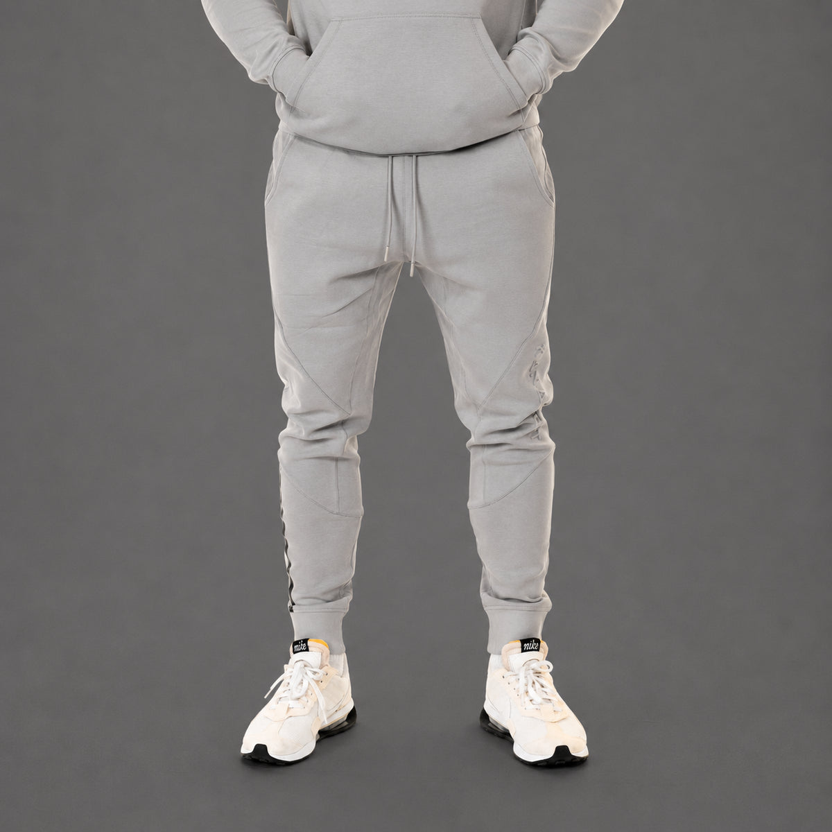 A person wearing a light gray matching sweatshirt and jogger pants set, shown from the chest down against a dark gray studio background. The person is wearing white sneakers.