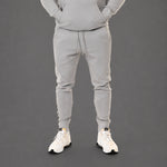 A person wearing a light gray matching sweatshirt and jogger pants set, shown from the chest down against a dark gray studio background. The person is wearing white sneakers.