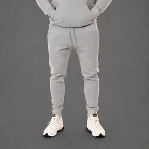 A person wearing a light gray matching sweatshirt and jogger pants set, shown from the chest down against a dark gray studio background. The person is wearing white sneakers.