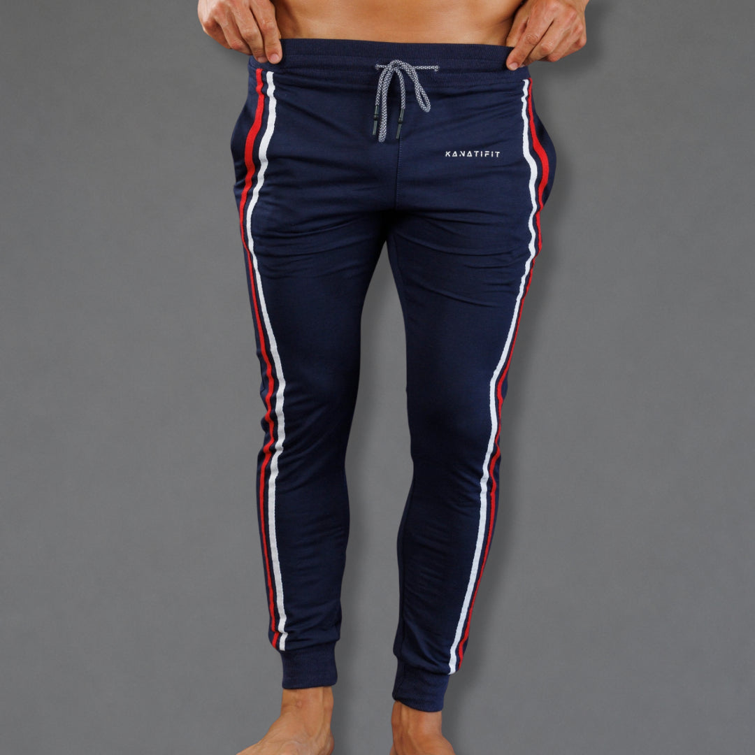 A person wearing navy track pants with red and white side stripes, shown from the waist down against a plain gray background. The pants have a drawstring waist and a small white 'KANSASFIT' logo on the right hip.