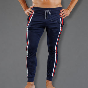 A man wearing navy blue track pants with red and white side stripes, standing against a solid gray background.