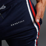 Close-up of navy athletic pants worn by a model, featuring a white KANATIFIT logo on the thigh and a red and white side stripe.