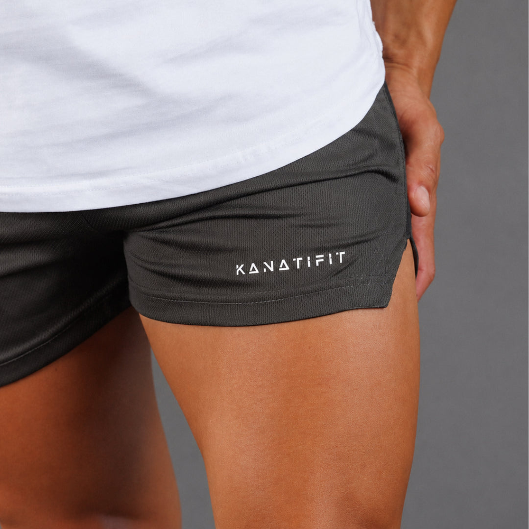 Close-up of a person wearing dark charcoal gray athletic shorts with the brand name 'KANATIFIT' printed in white on the left leg, paired with a white t-shirt against a solid gray background.