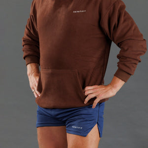 A person wearing a brown long-sleeve sweatshirt and blue athletic shorts, shown from the chest down against a plain gray studio background. Small white 'KANATIFIT' branding is visible on both garments.