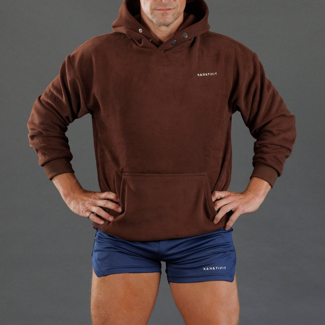 A man wearing a brown pullover hoodie with a small white KANATIFIT logo on the left chest, paired with navy athletic shorts against a solid gray studio background.