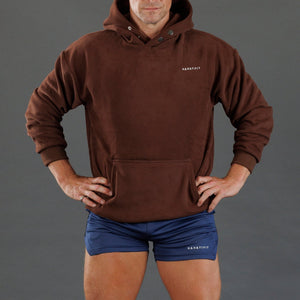 A man wearing a brown pullover hoodie with a small white KANATIFIT logo on the left chest, paired with navy athletic shorts against a solid gray studio background.