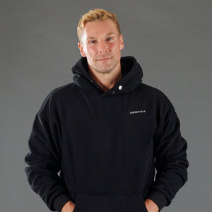 A man wearing a black pullover hoodie with a small white 'KANATIFIT' logo on the left chest, photographed against a solid gray studio background.