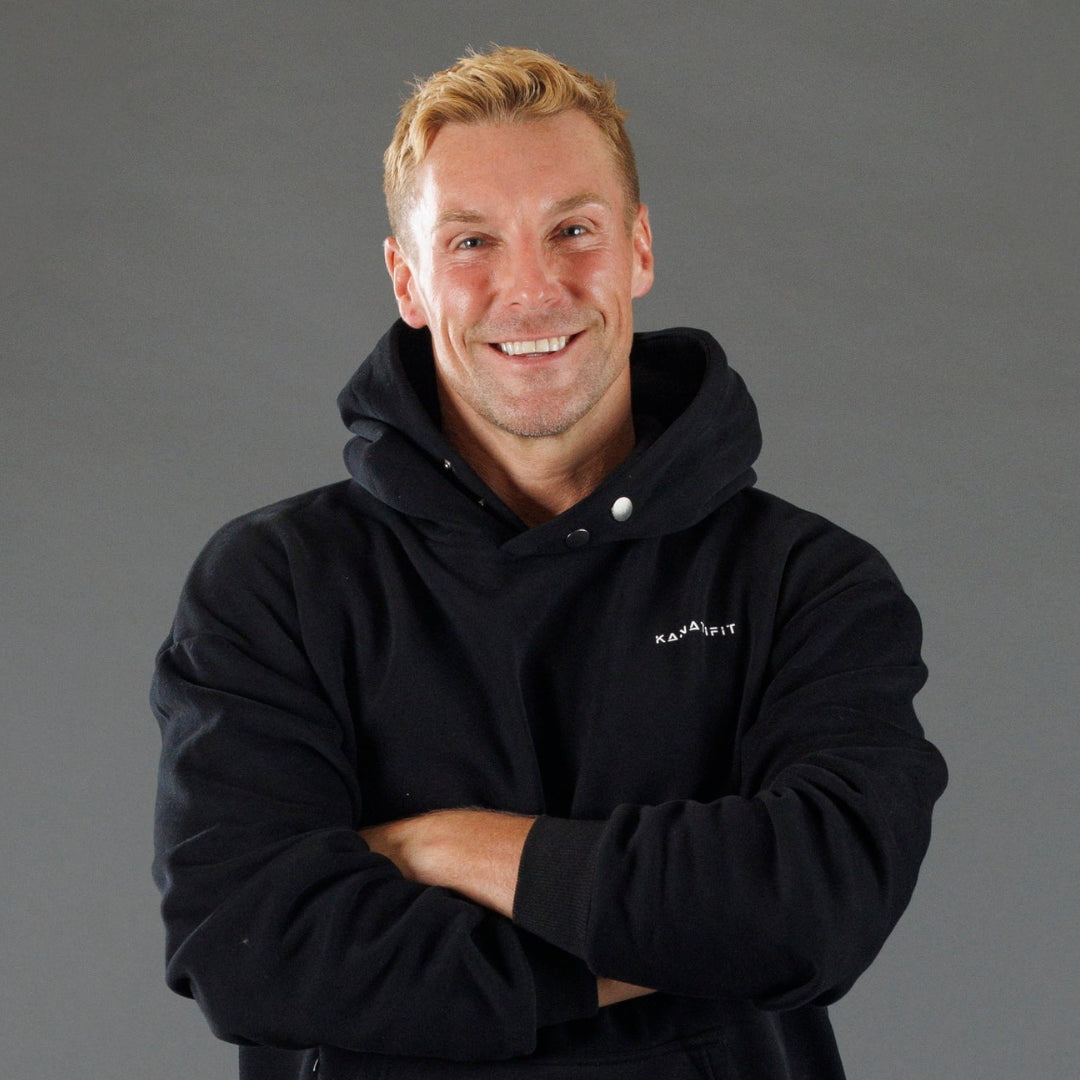 A smiling man with blond hair wearing a black pullover hoodie with a small white 'KANATIFIT' logo on the left chest, posed with arms crossed against a solid gray studio background.