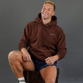 A smiling man wearing a brown pullover hoodie with a small white 'KANATIFIT' logo on the chest, paired with navy athletic shorts, sitting on a dark trunk against a solid gray studio background.
