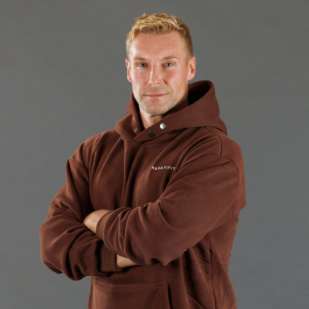 A man wearing a brown hooded sweatshirt with 'KANATIFIT' printed in white on the left chest, posed with arms crossed against a solid gray studio background.